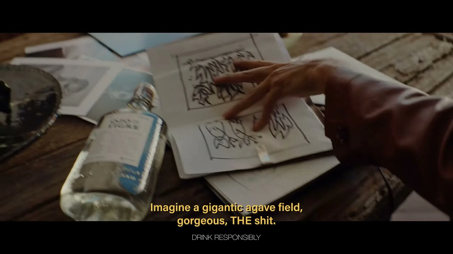 A man in a colorful shirt sits on a chair, gesturing with his hands as he speaks, while a bottle of Ojo de Tigre Mezcal and a sketchbook are on a wooden table.