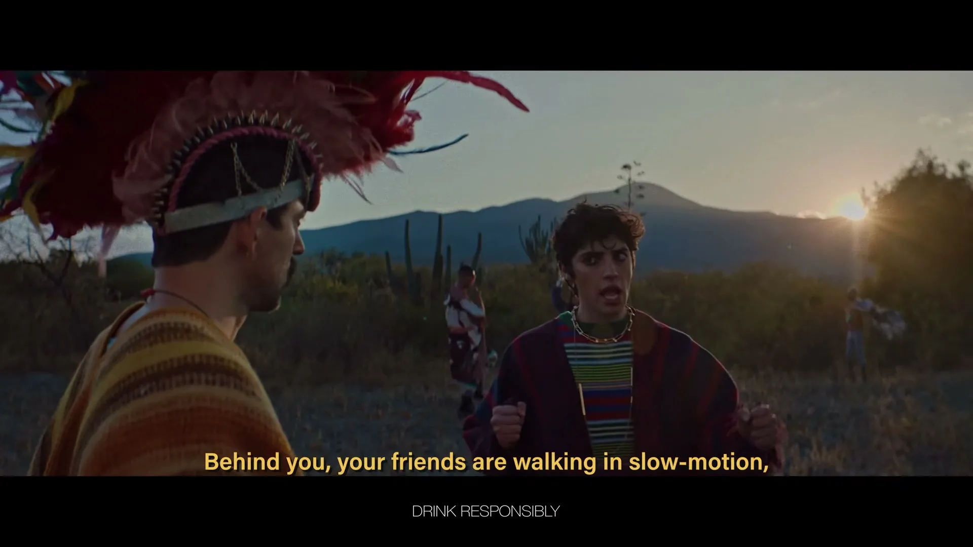 Two men in traditional attire stand in a desert landscape at sunset, one speaking animatedly to the other, with more people in the background.