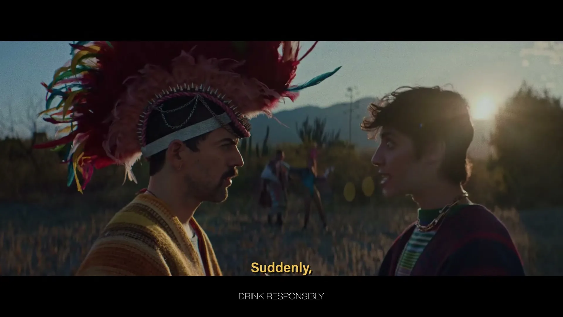 Two men in traditional Mexican attire, one with a large feathered headdress, stand facing each other in a field at sunset, talking intently.
