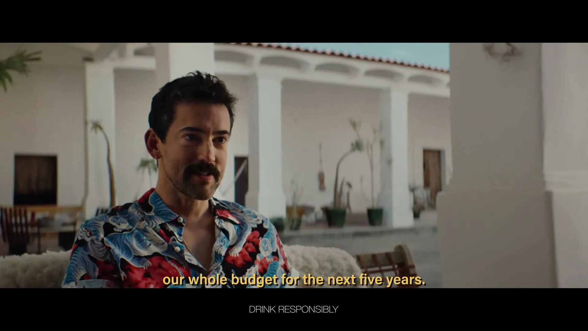 A man with a mustache wearing a colorful shirt sits on a couch, looking slightly to the right, in an outdoor patio setting.