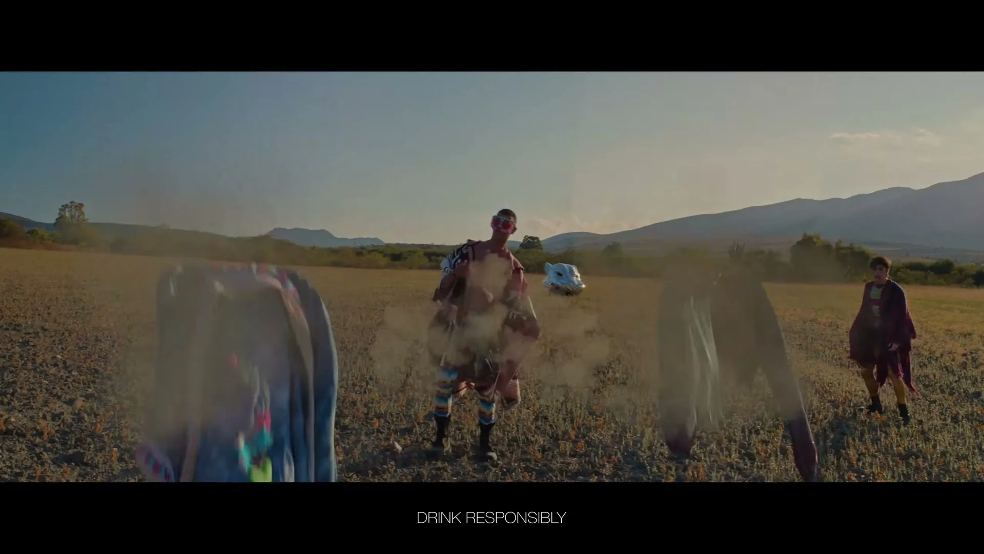 Four people in colorful, eclectic outfits stand in a dry, open field, then their clothes explode off them, leaving them naked.
