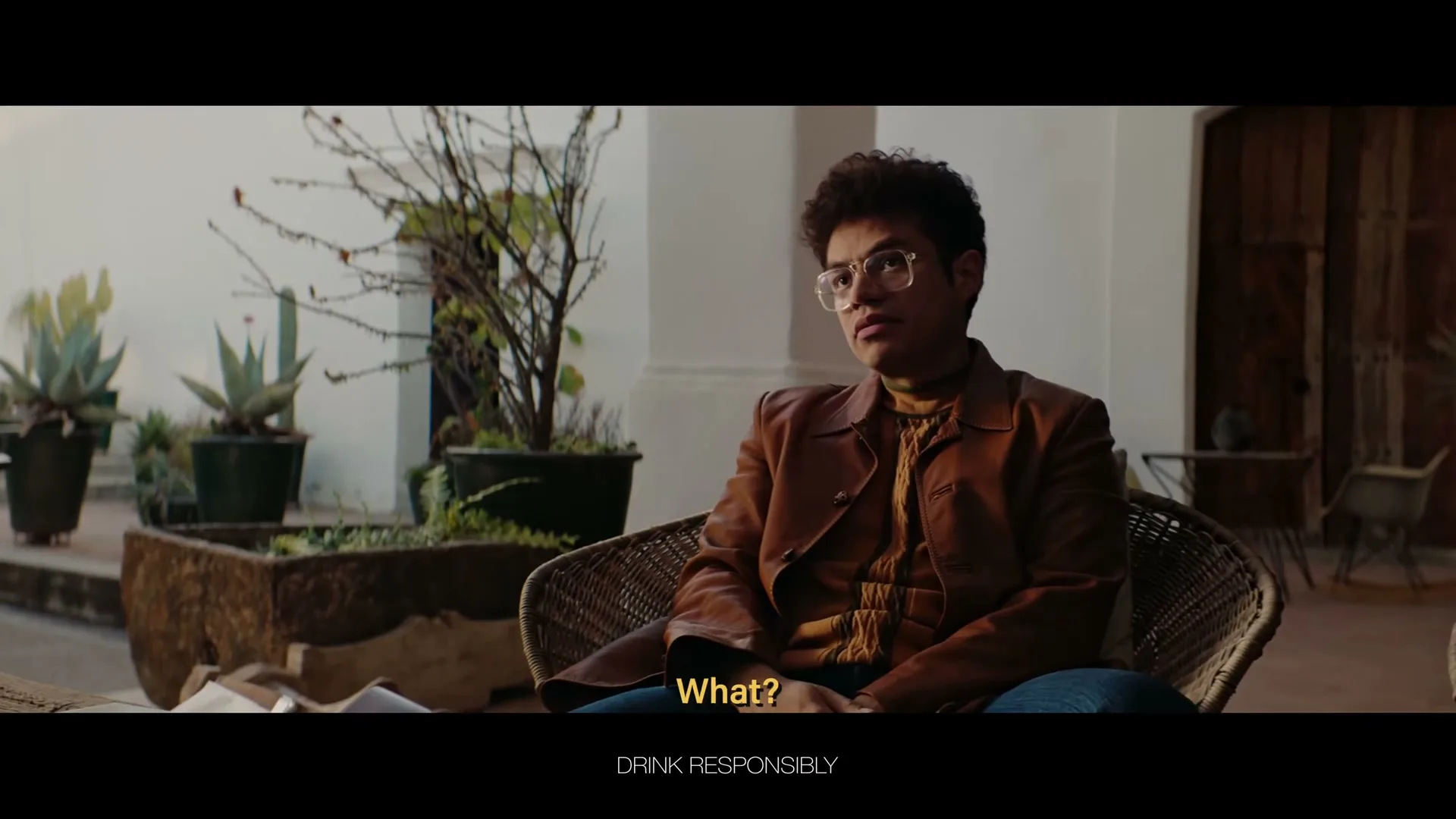 A young man with curly hair and glasses sits in a wicker chair, looking slightly confused, in an outdoor patio with potted plants.