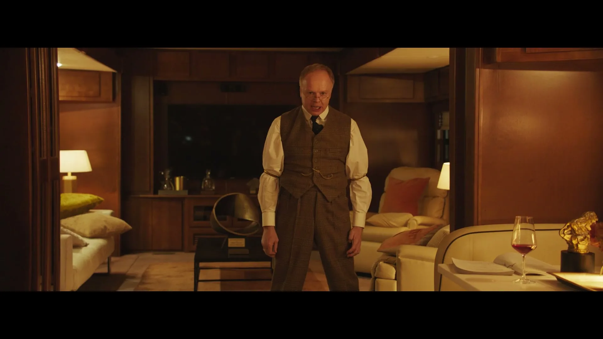 A man in a brown suit and white shirt stands in a dimly lit, wood-paneled room, looking around with a confused expression.