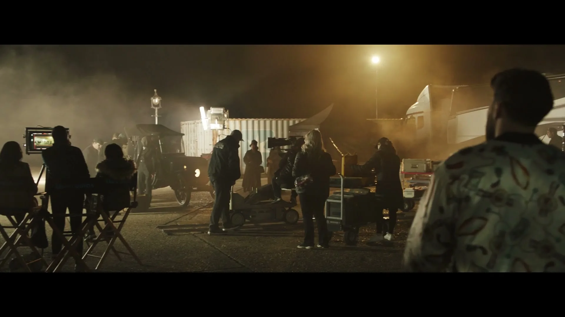 A film crew works at night on a set, with a vintage car and various equipment visible under bright artificial lights and smoke.