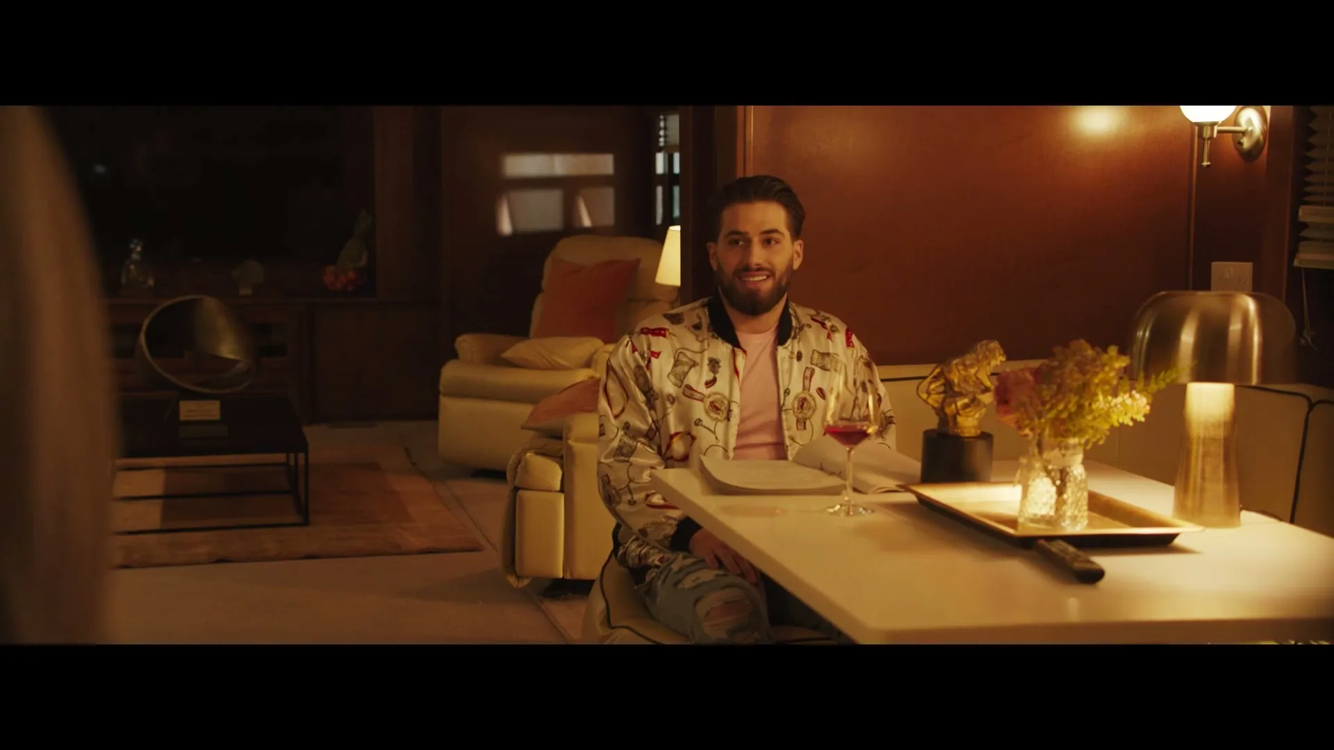A man with a beard and a patterned jacket sits at a table, smiling and looking towards the camera in a dimly lit living room.