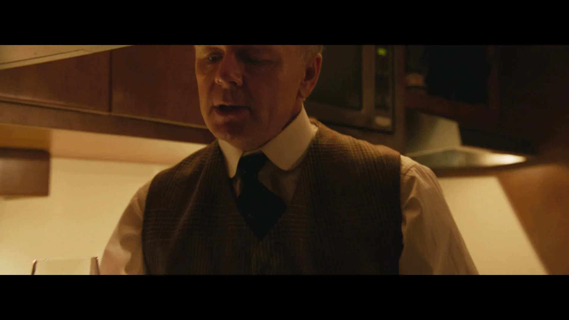 A man in his 50s, dressed in a white shirt and brown vest, is seen from the chest up, speaking in a dimly lit kitchen.