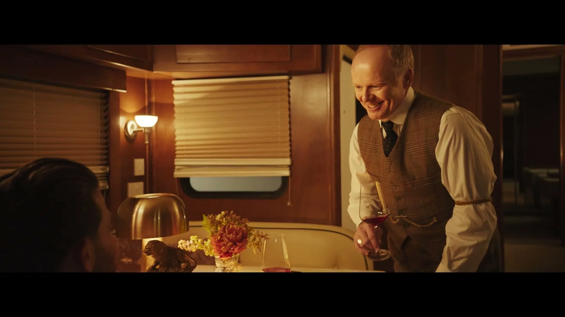 A man in a tweed vest and white shirt holds a glass of red wine, smiling as he talks to another man across a table in a luxurious train car.