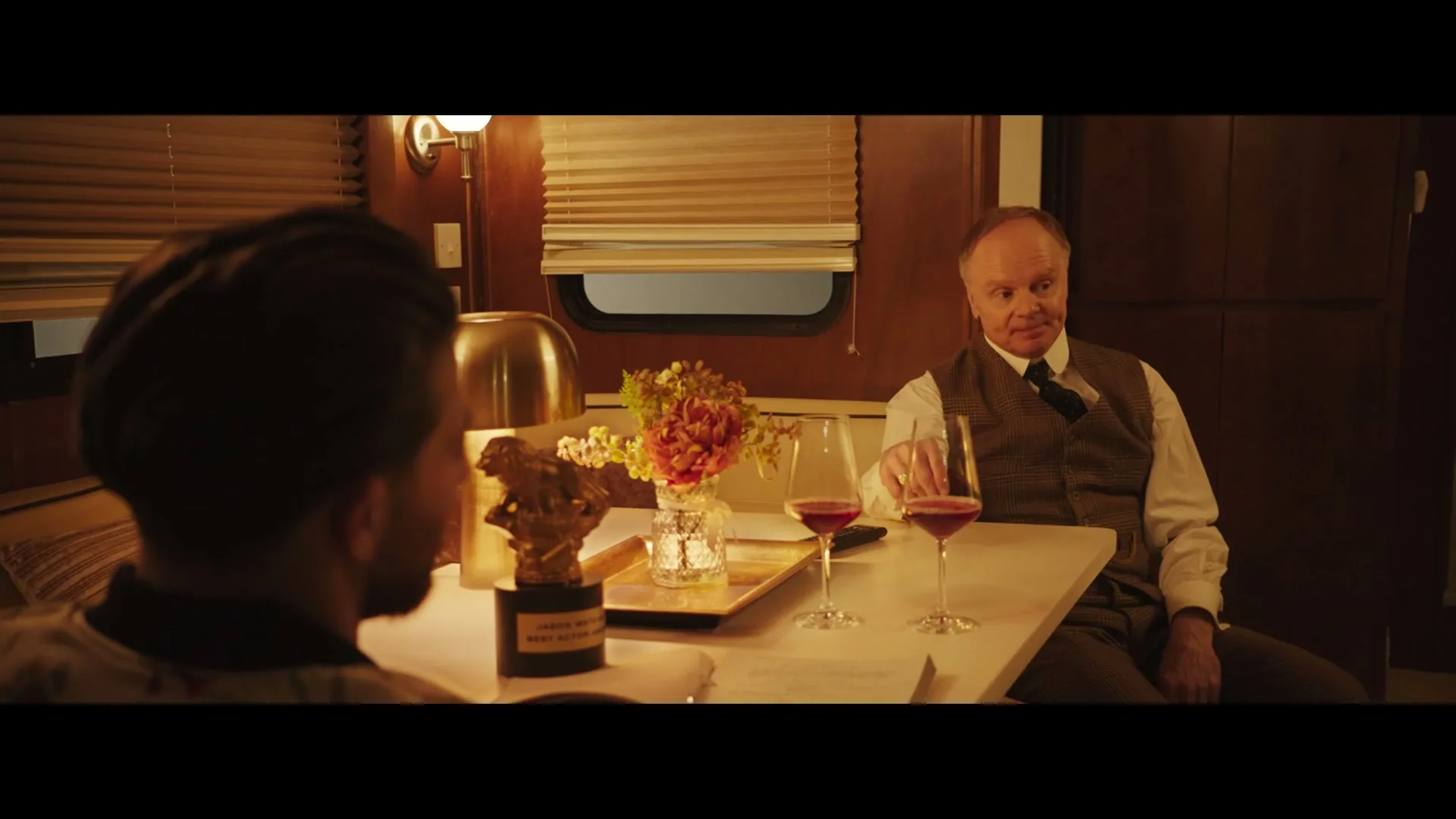 Two men are sitting at a table in a luxurious train car, with one man in a vest and tie smiling at the other, who is seen from behind.