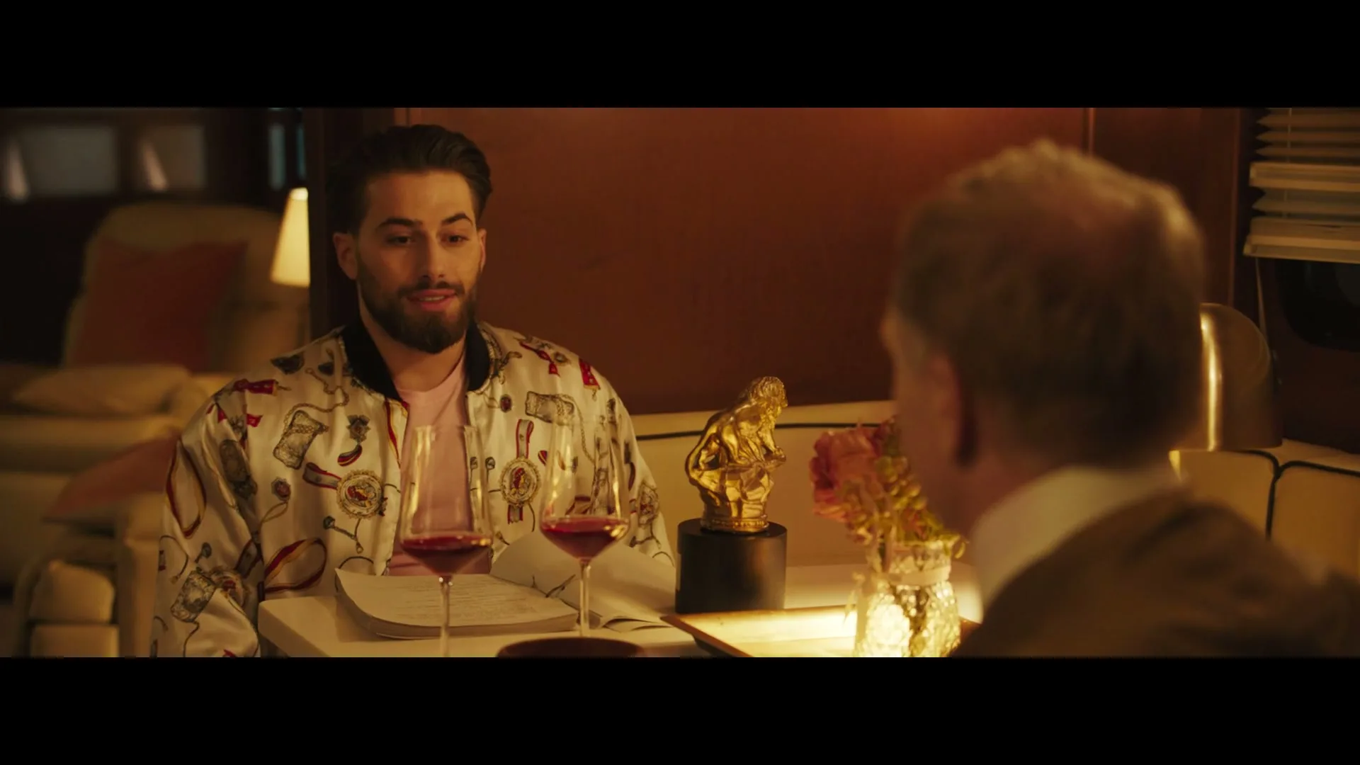 A young man with a beard, wearing a patterned jacket, sits at a table with wine glasses, talking to an older man in a dimly lit, luxurious interior.