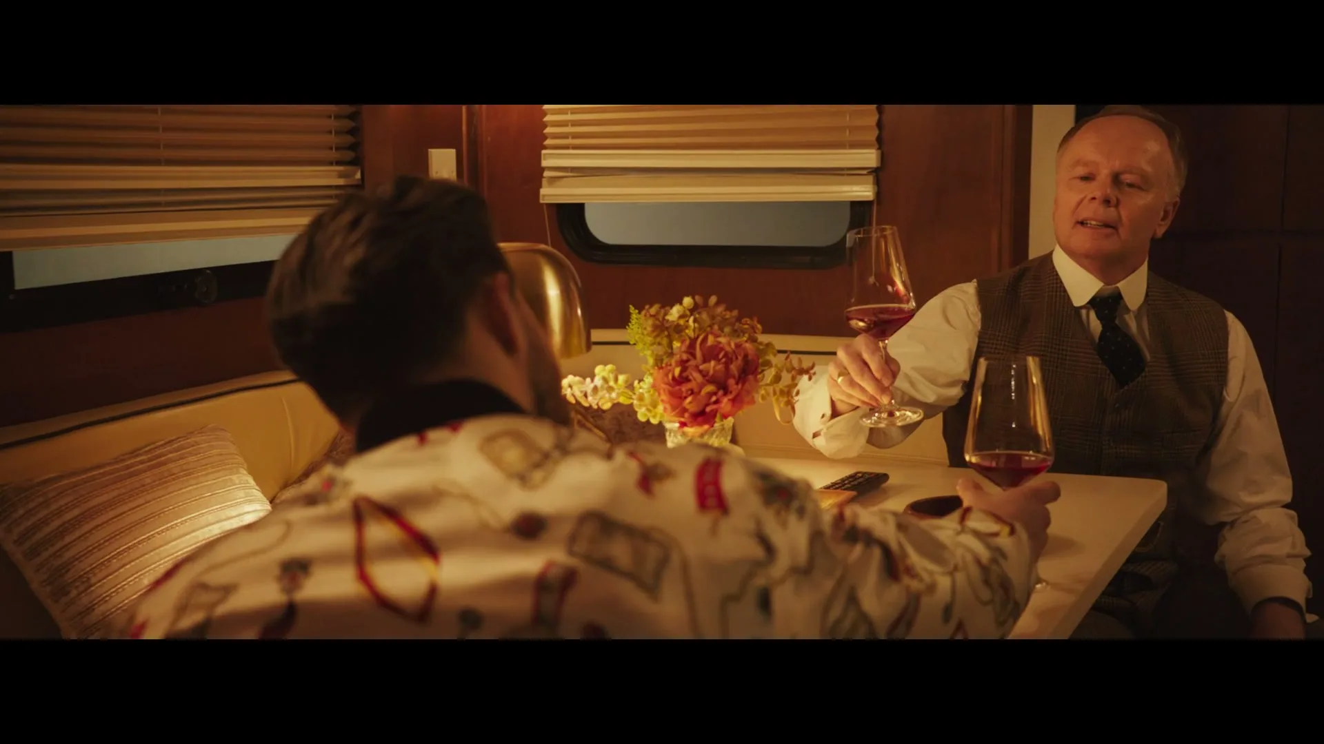 Two men, one with a patterned shirt and the other in a vest, sit at a table with wine glasses and a trophy, toasting each other in a cozy, wood-paneled room.