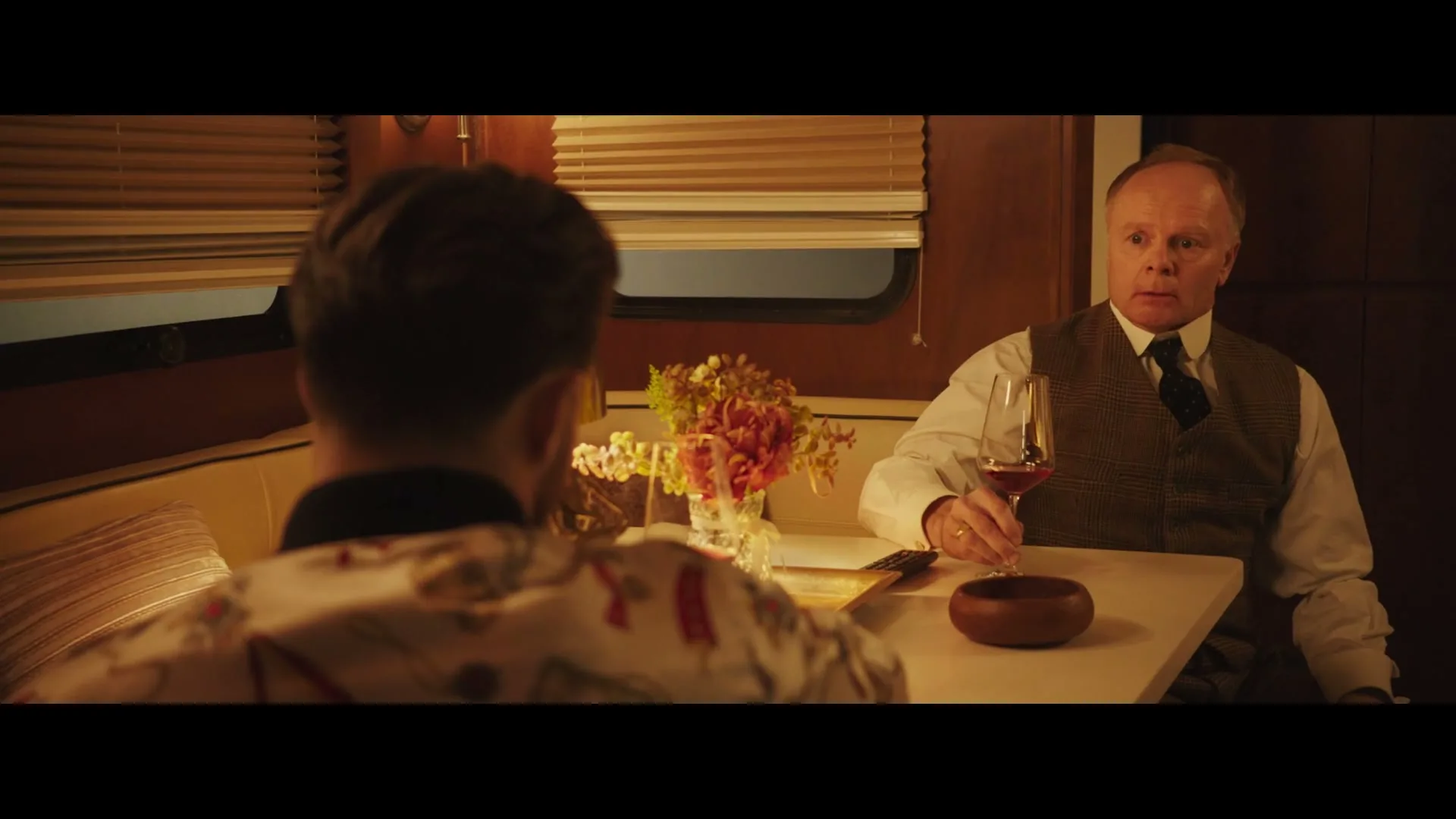 Two men sit opposite each other at a table in a train car, one holding a glass of red wine, the other with his back to the camera.