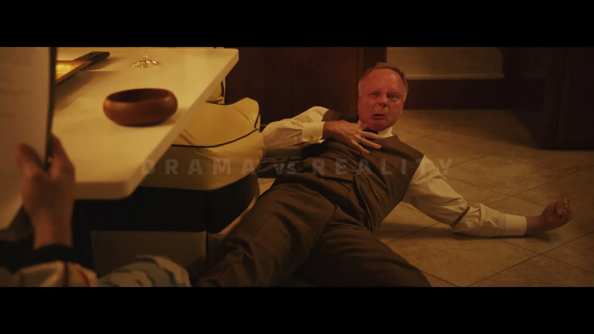 A man in a brown suit and white shirt lies on the floor, clutching his chest, with a distressed expression, while text overlays the scene.