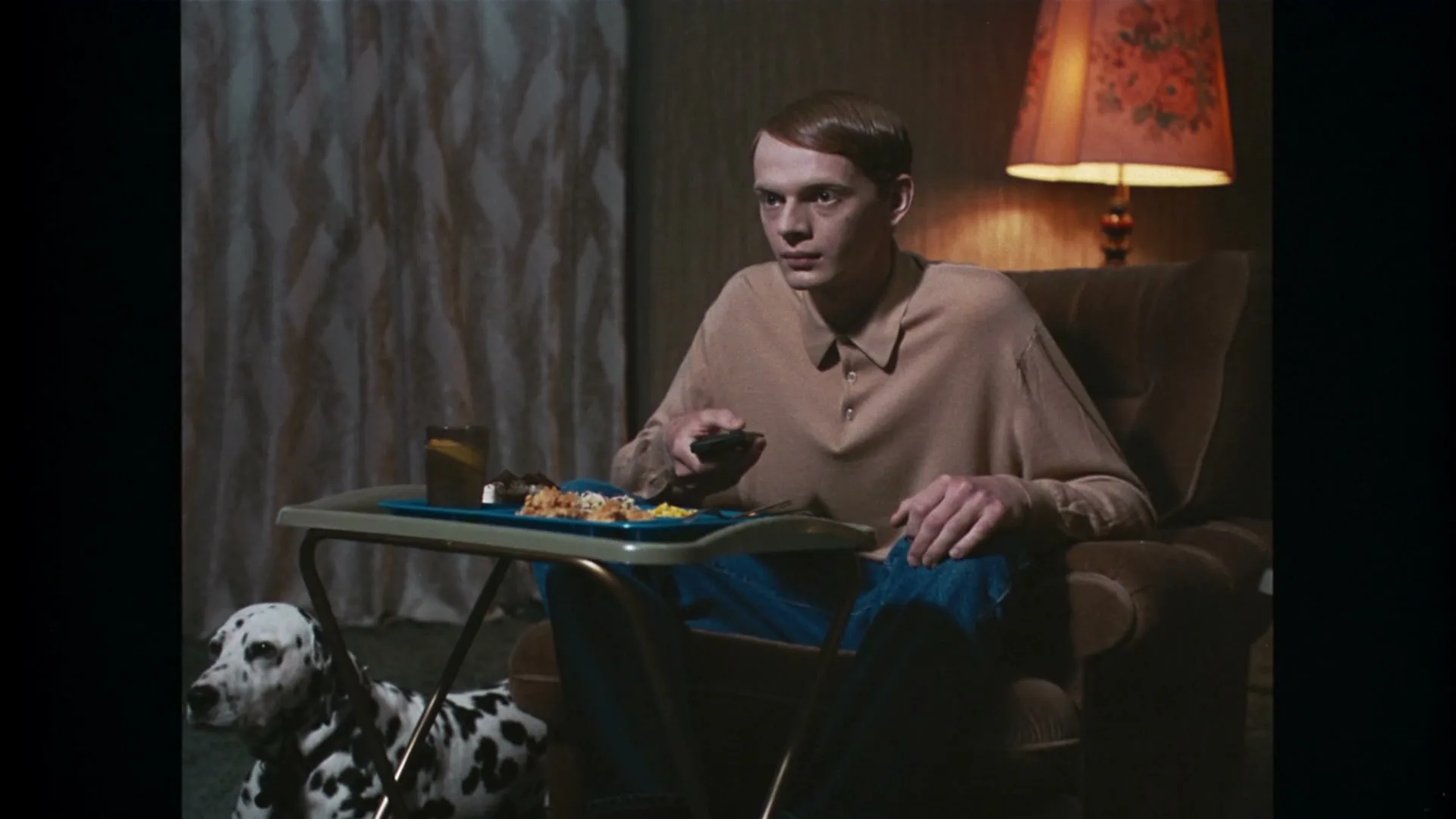 A young man with a bowl cut sits in a recliner, holding a remote, with a TV dinner on a tray and a dalmatian dog beside him in a dimly lit living room.