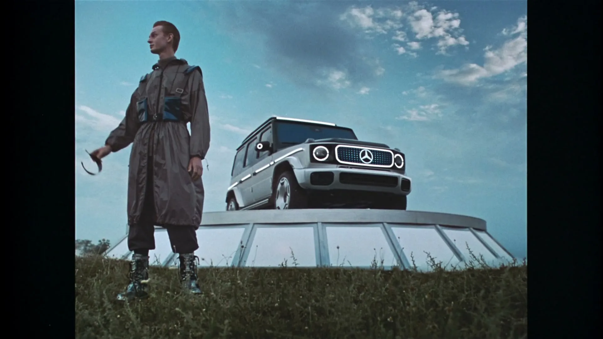 A young man in futuristic attire stands next to a Mercedes-Benz G-Wagen on a UFO-like platform in a grassy field under a cloudy sky.