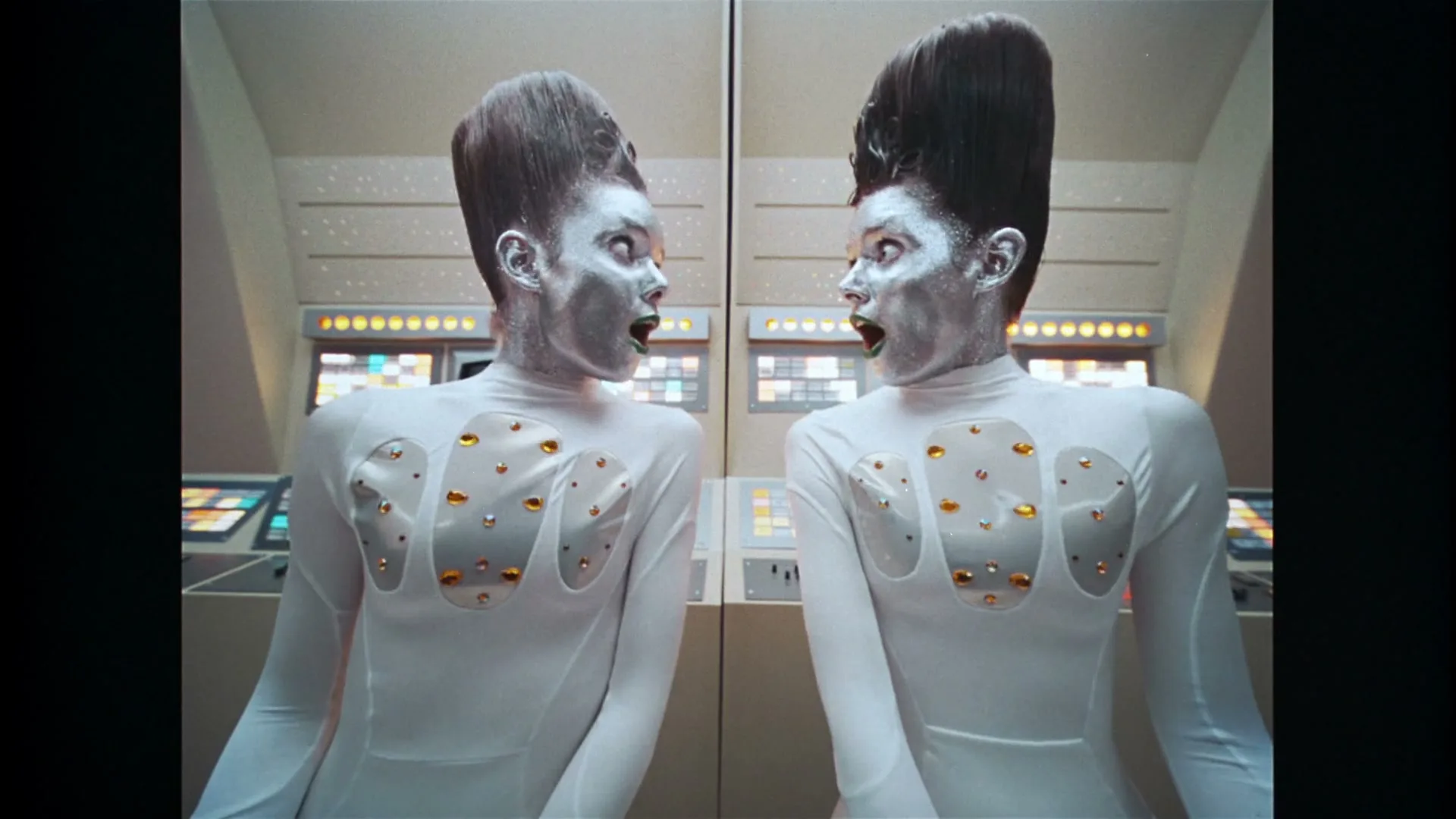 Two women with silver faces, green lipstick, and tall hair, wearing white futuristic bodysuits with clear cutouts, look at each other in a spaceship.