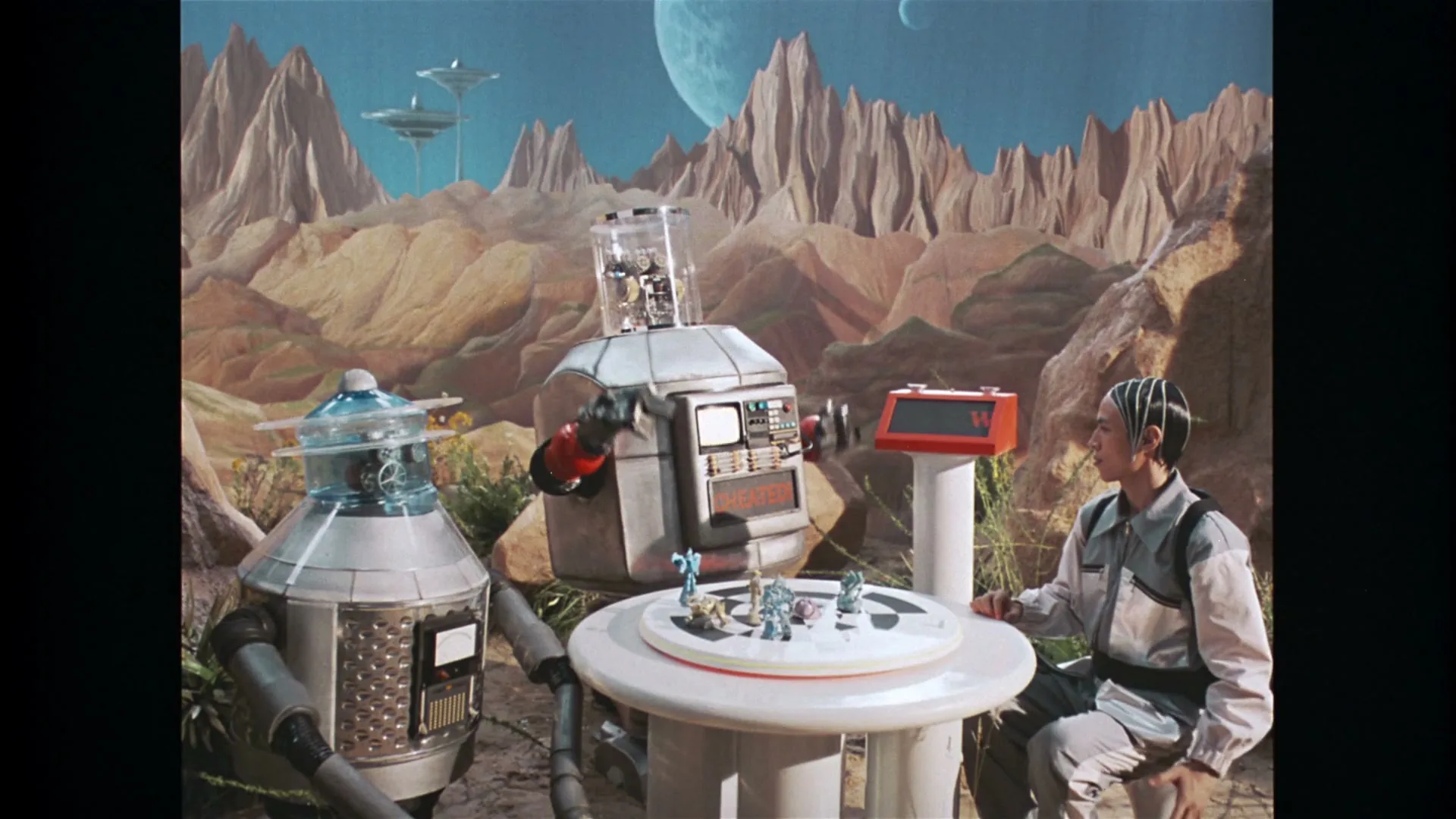 A young man with braided hair sits at a table with two robots on an alien planet, playing a board game, as one robot shoots a green laser.