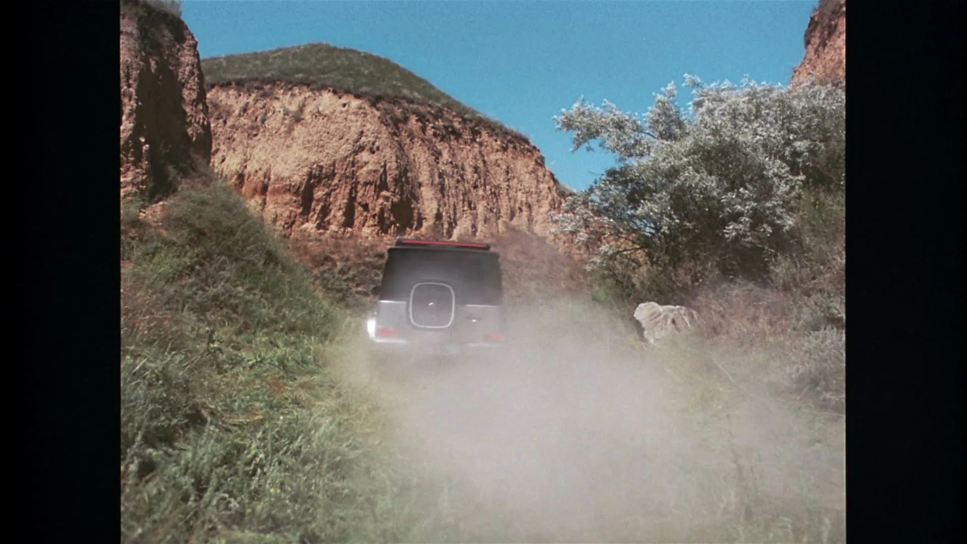 A silver Mercedes-Benz G-Wagen drives away from the camera on a dirt path, kicking up dust between two grassy hills under a clear blue sky.