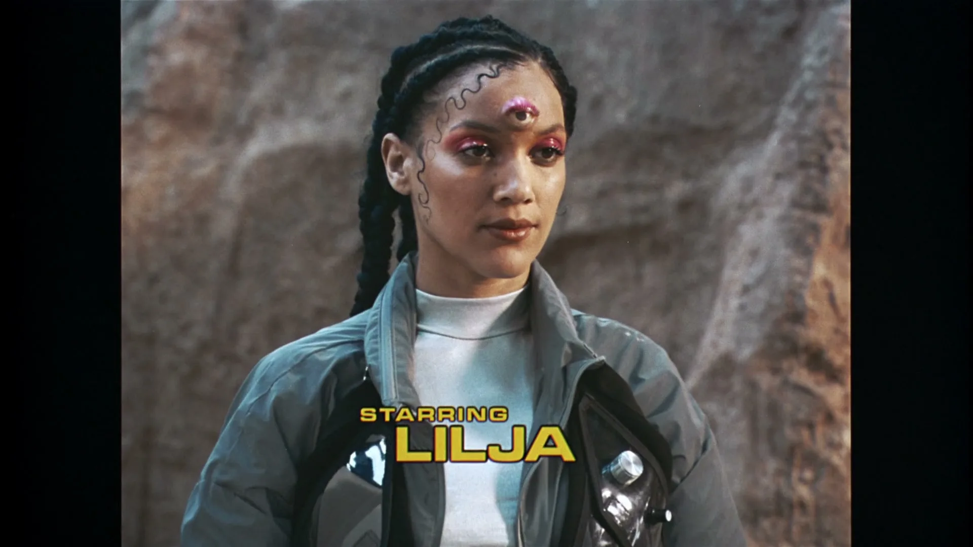 A woman with braided hair and a third eye on her forehead looks through futuristic binoculars, then lowers them, revealing her face and the text "STARRING LILJA" on screen.