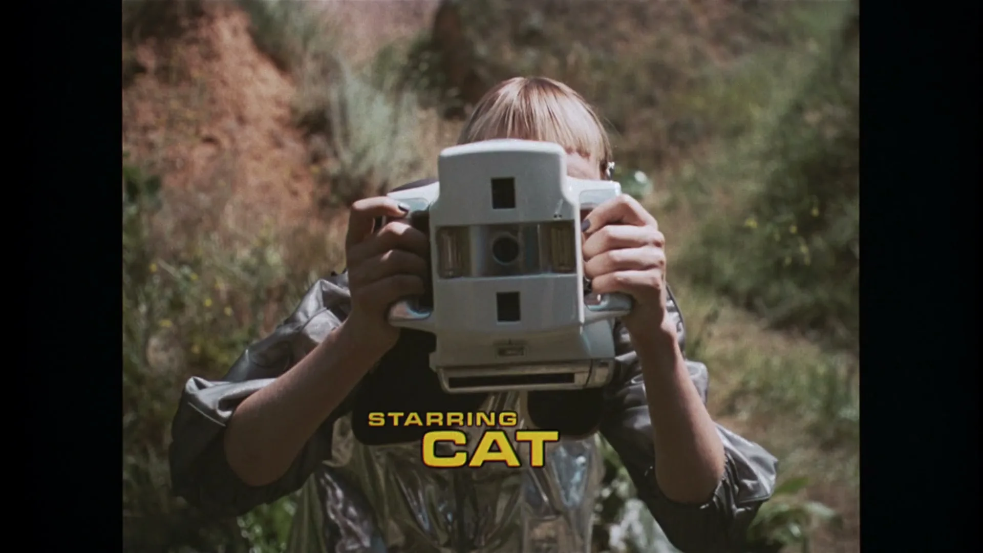 A blonde person in a futuristic silver and black outfit holds up a white camera, obscuring their face, in a rocky, grassy outdoor setting.