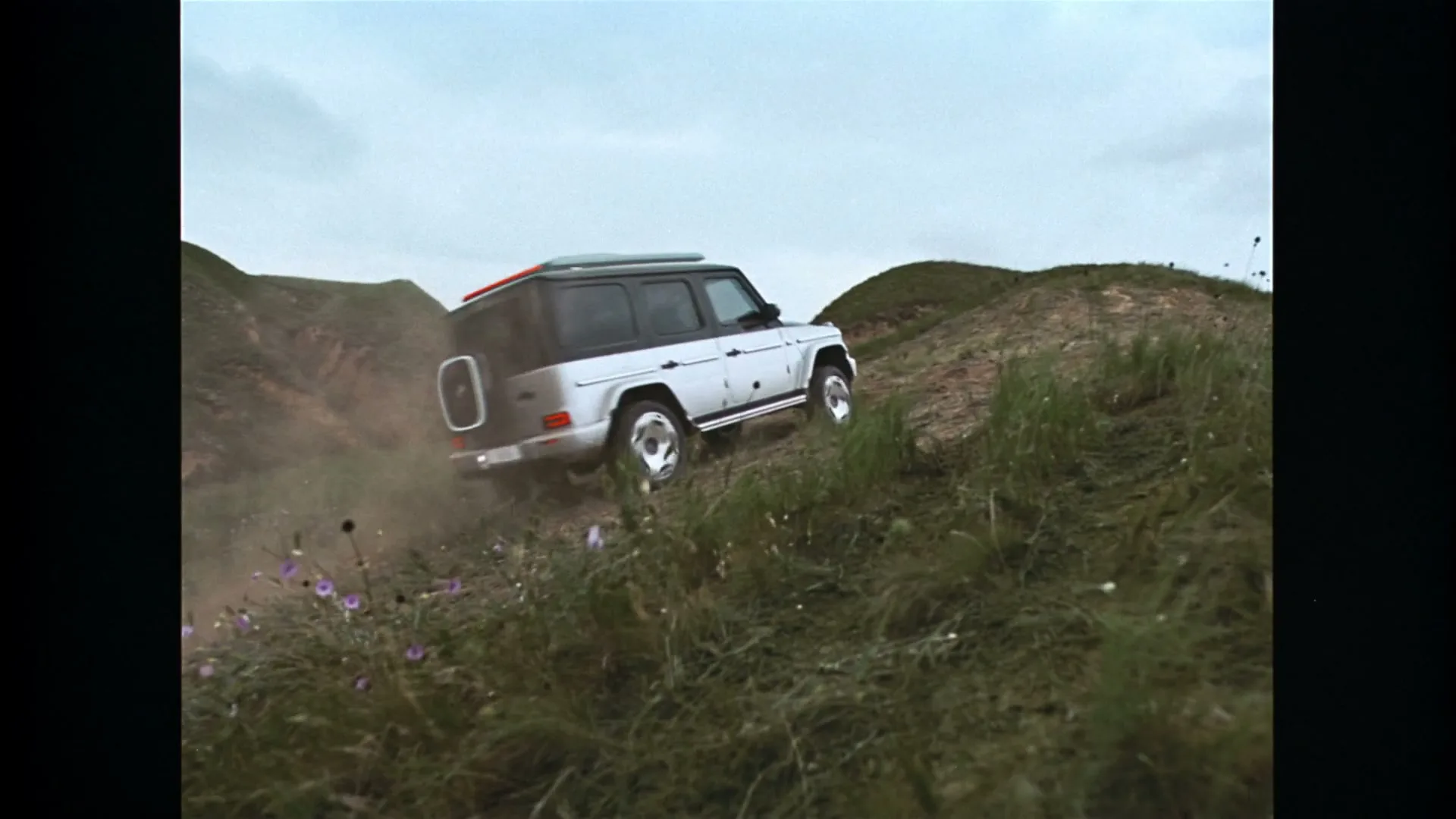 A white Mercedes-Benz G-Wagen drives up a steep, grassy hill, kicking up dust and dirt, under an overcast sky.