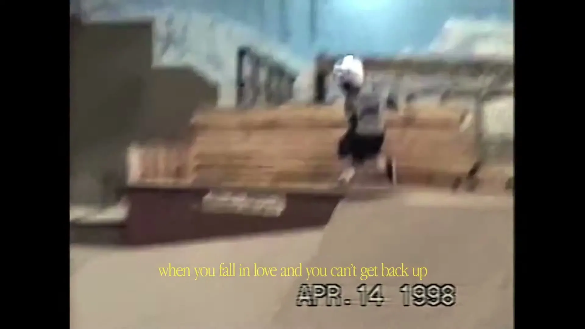 A young boy in a helmet and pads attempts to skateboard on a ramp in an indoor skatepark, captured in archival footage.