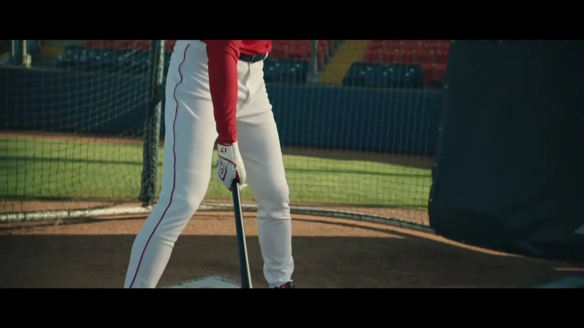 A baseball player in a red shirt and white pants holds a bat, preparing to swing in a batting cage at a stadium.