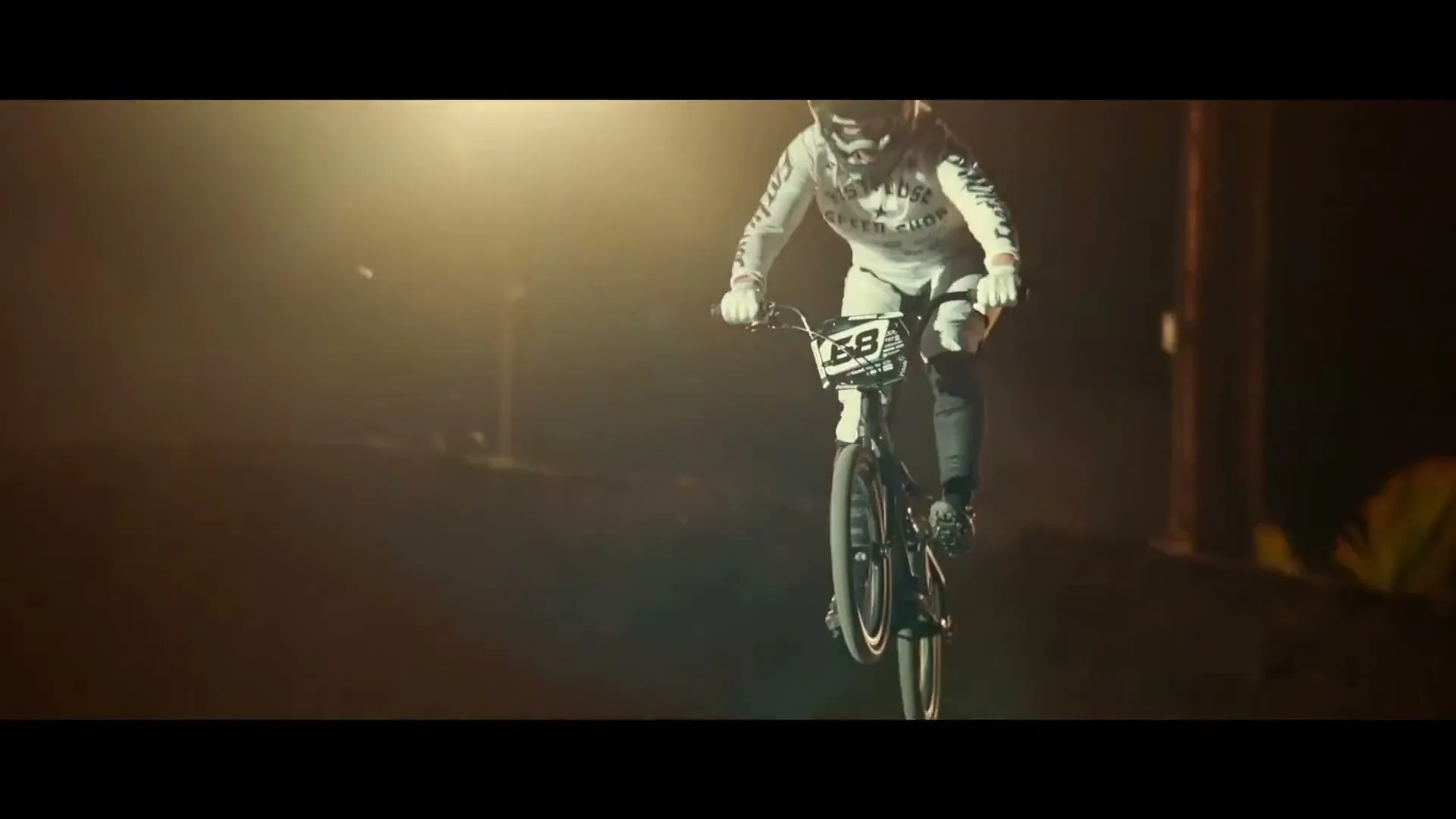A BMX rider in a white uniform and helmet rides a bike on a dirt track at night, illuminated by a bright light.