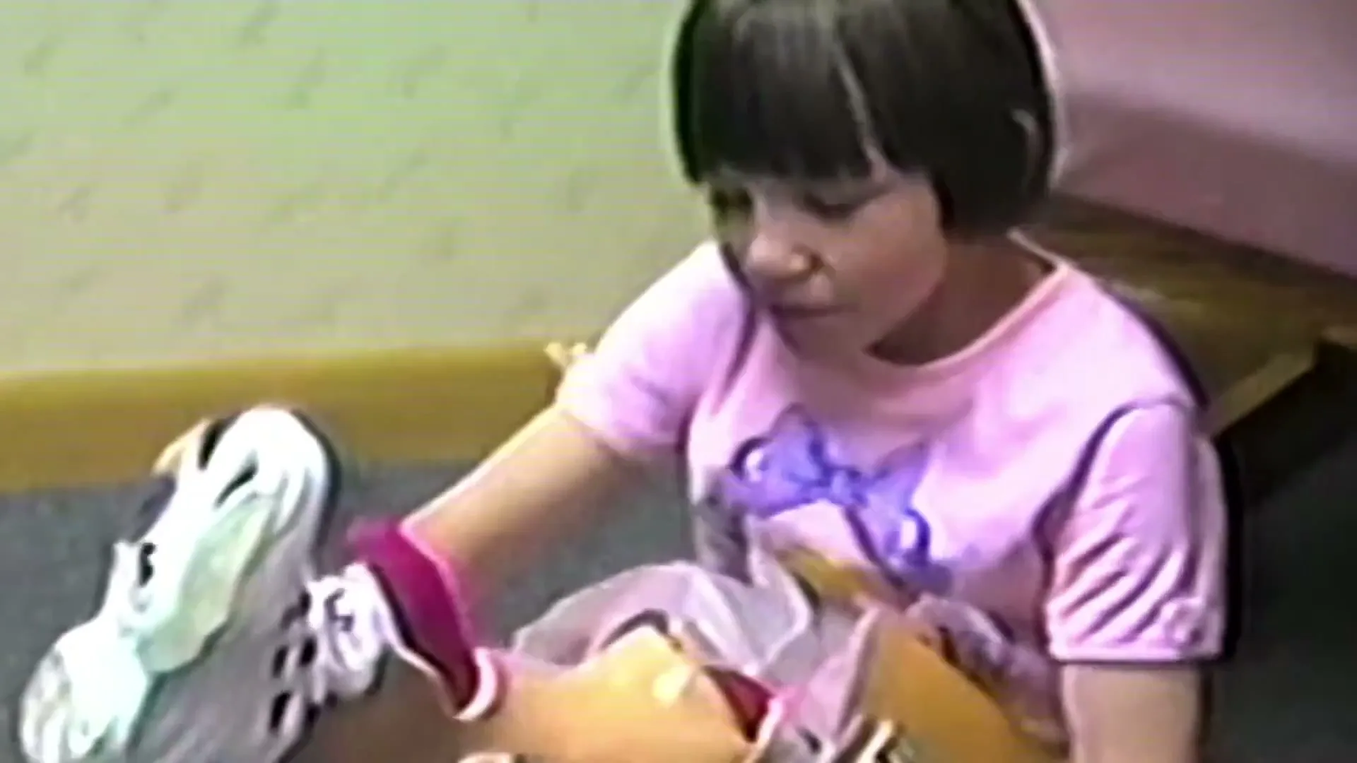 A young girl with short dark hair and a pink t-shirt sits on the floor, playing with a doll and a white shoe in an interior setting.