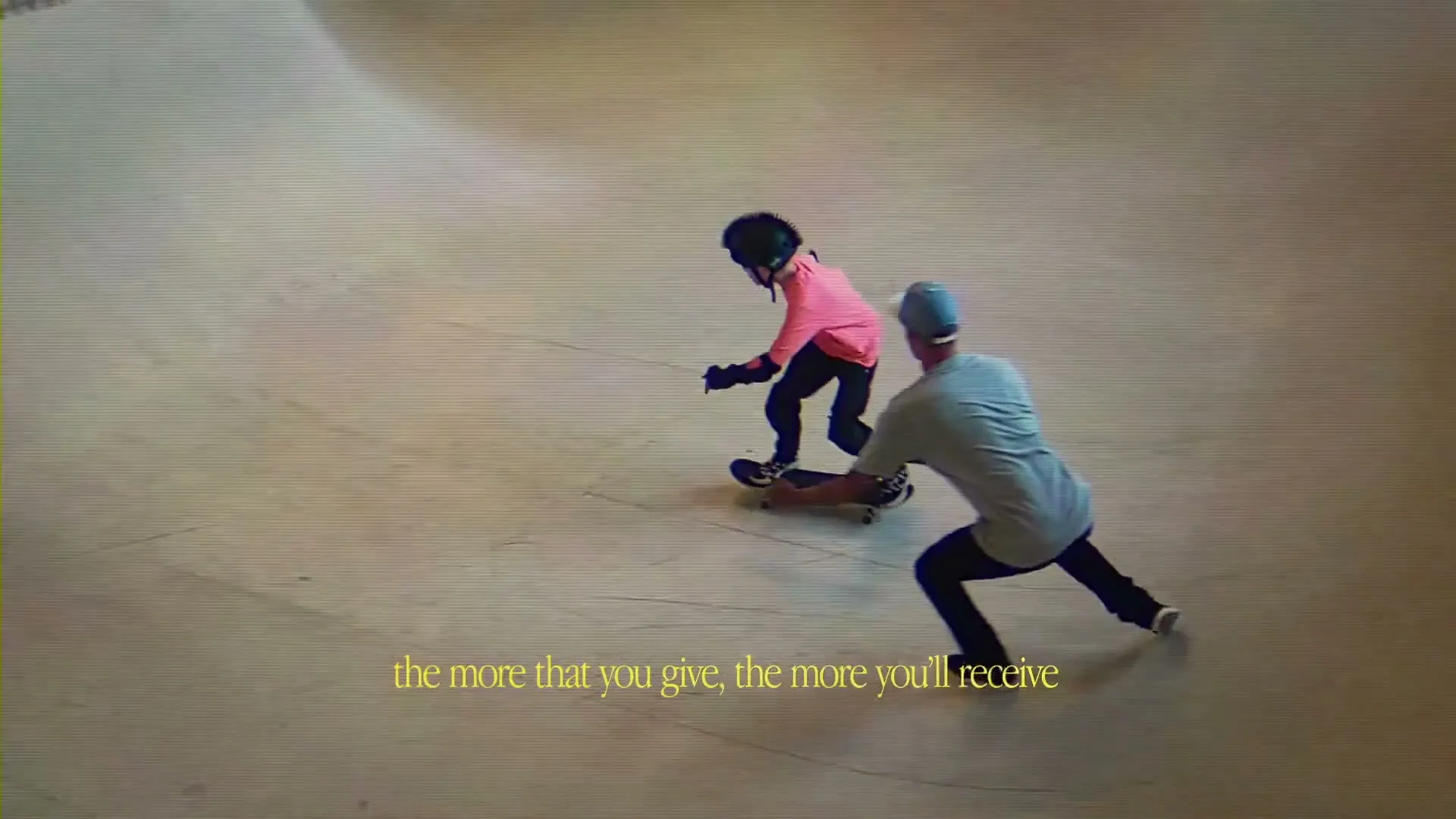 A young boy in a helmet and pads skates down a ramp in a skatepark, while an adult male reaches out to help him.