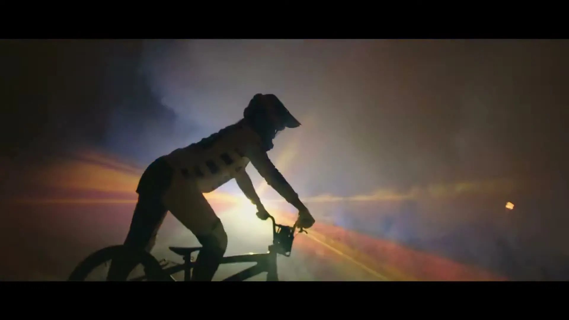 A BMX rider in a helmet and full gear is silhouetted against a smoky, brightly lit background, preparing to ride.