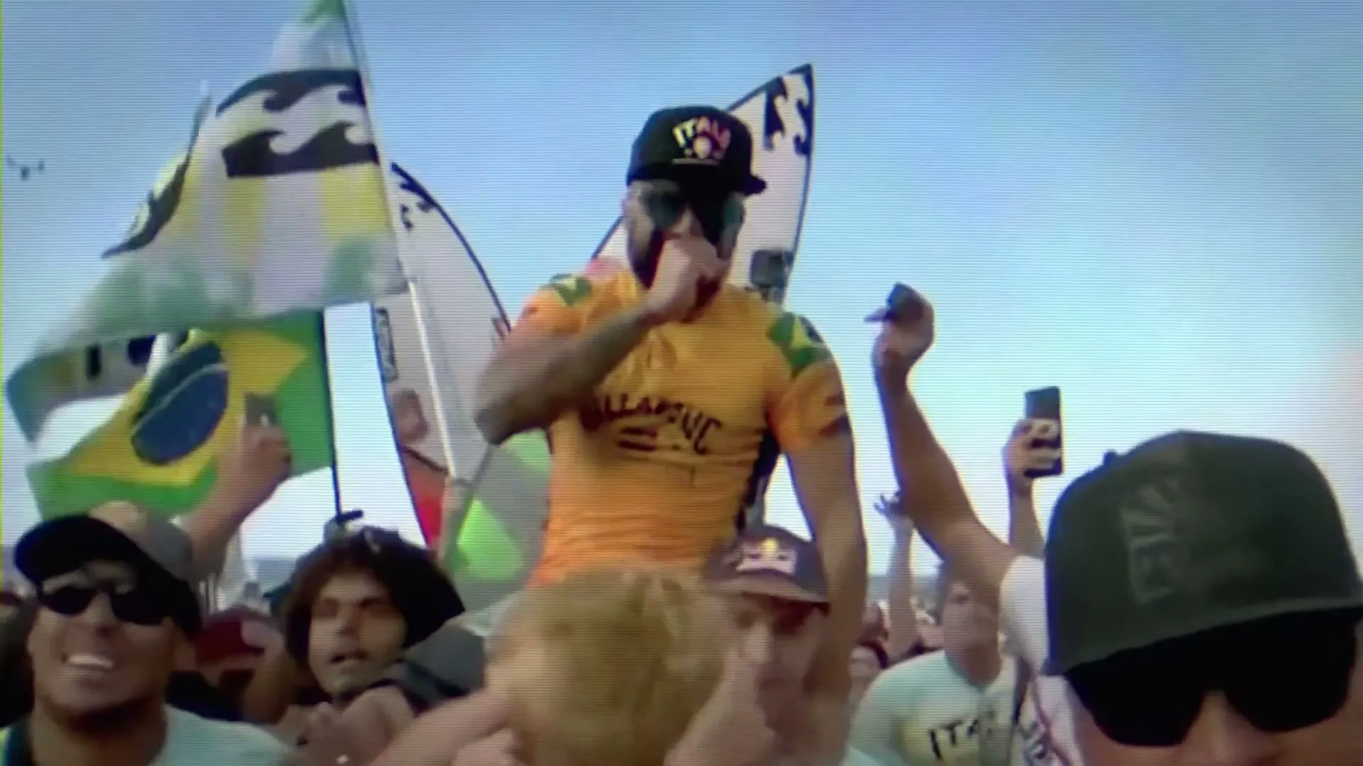 A male surfer, wearing a yellow shirt and black cap, is carried on the shoulders of a crowd, celebrating his victory on a beach.