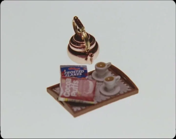 A miniature breakfast tray with a kettle, two cups, and two cereal boxes, one of which is placed on top of the other, is shown on a white background.