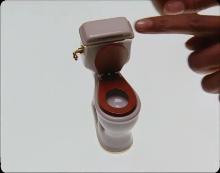 A miniature toilet with a brown seat is shown on a white background, as a finger reaches in to close the lid.