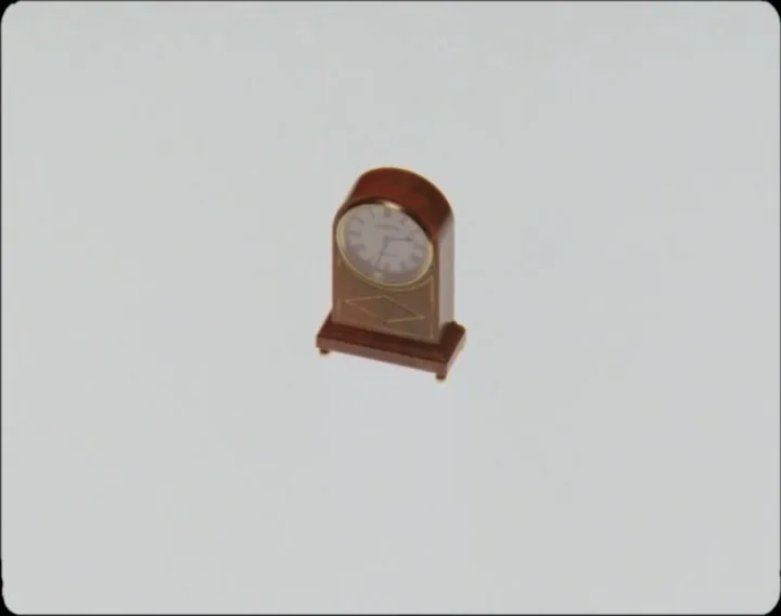 A finger pushes a small, brown, old-fashioned clock off-screen, leaving a plain white background.