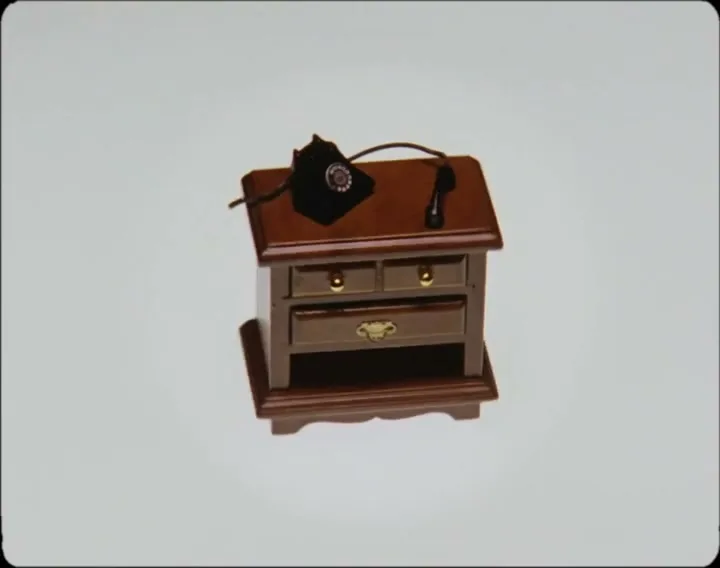 A miniature wooden nightstand with a black rotary phone on top rotates slowly against a plain white background.