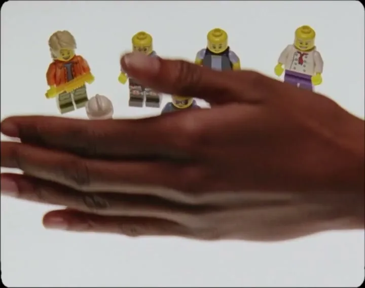 A dark-skinned hand sweeps across a white surface, clearing away a collection of Lego minifigures and accessories.