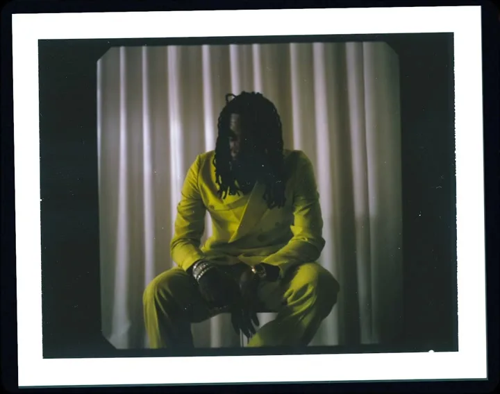 A man with dreadlocks, wearing a bright yellow suit, sits with his head down in a dimly lit room with white curtains.