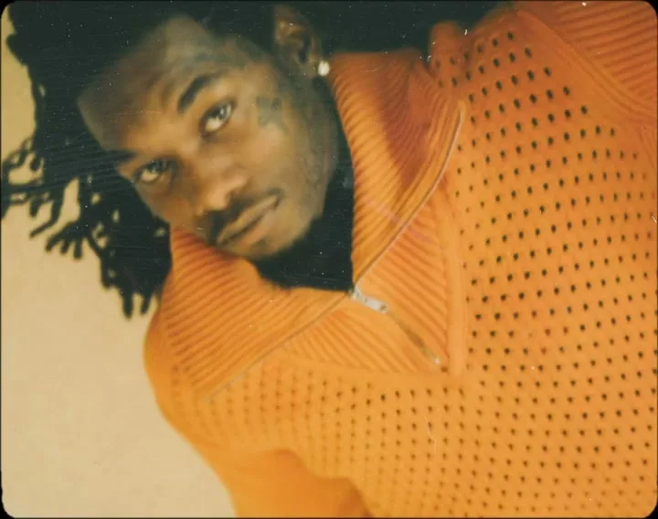 A man with dreadlocks and tattoos on his face looks directly at the camera, wearing an orange knitted sweater in a studio.