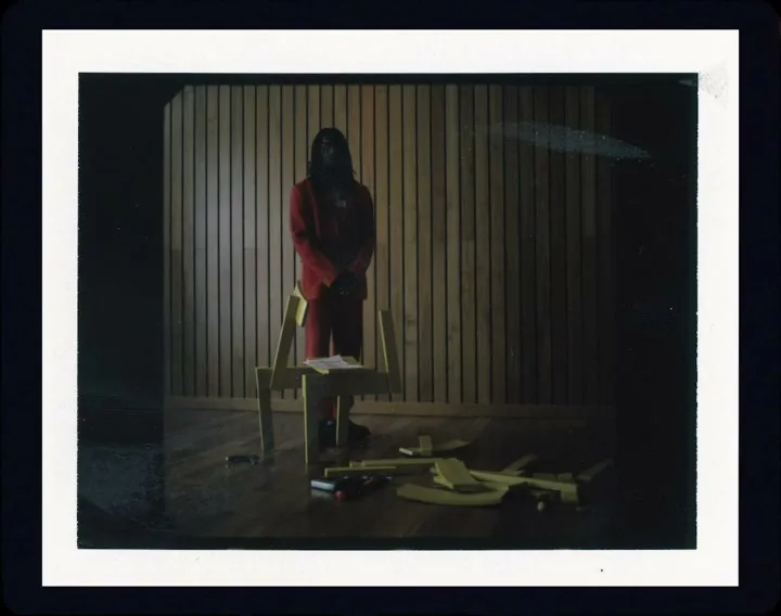 A person in a red suit stands behind a partially assembled yellow chair, with more chair parts scattered on the wooden floor in a dimly lit room.