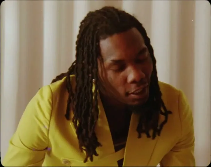 A Black man with dreadlocks in a yellow blazer speaks directly to the camera in a studio with a white pleated background.
