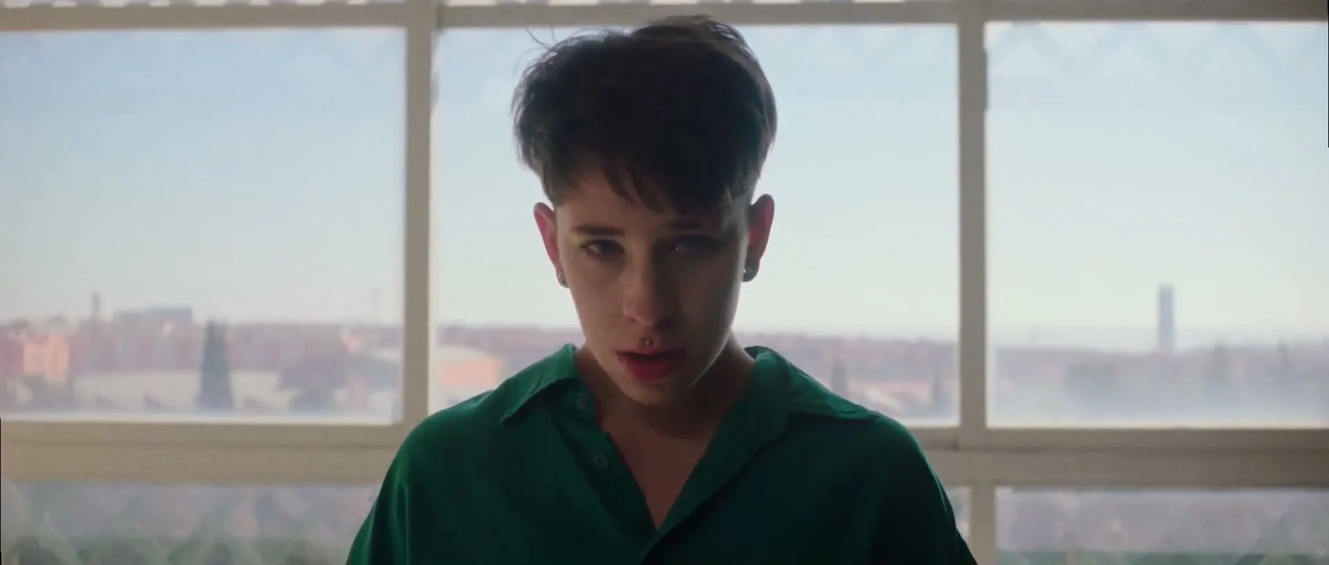 A young person with short dark hair and a green shirt looks directly at the camera with a serious expression, standing in front of a large window overlooking a city.