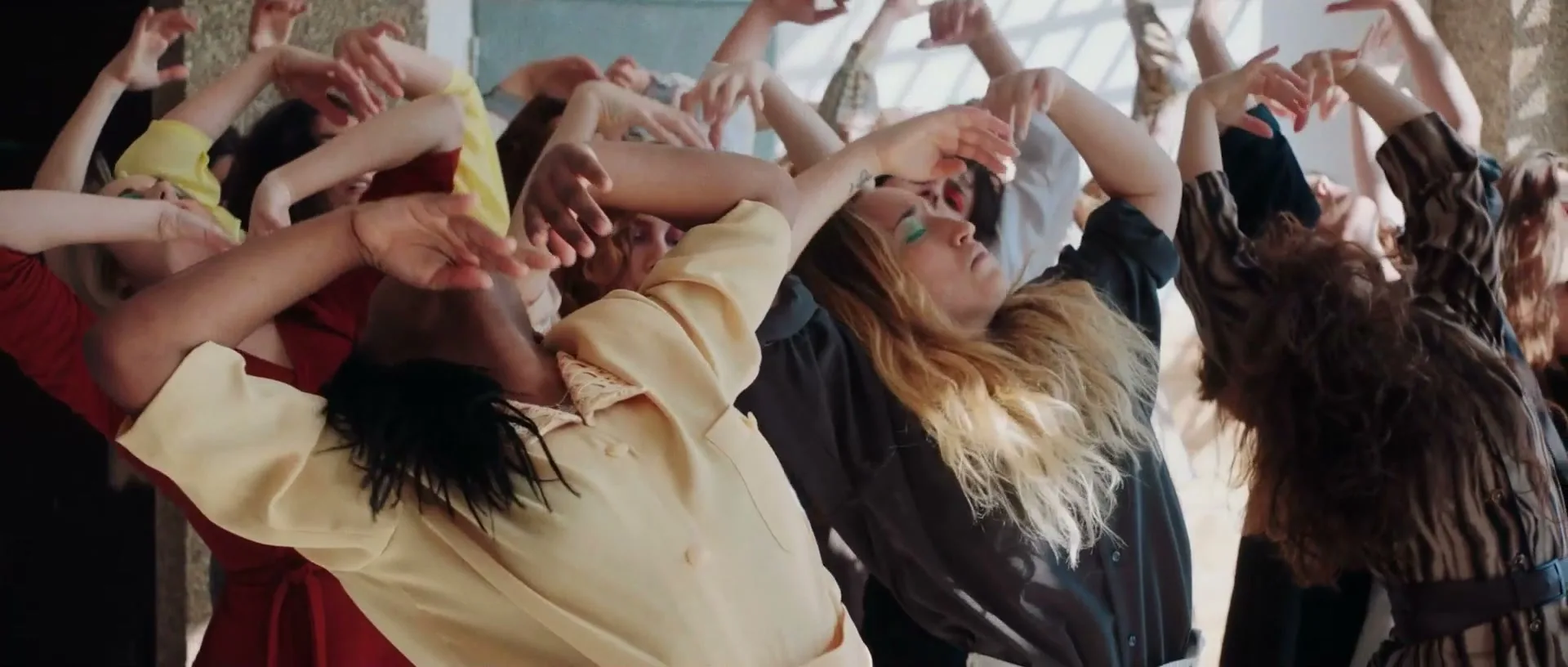 A diverse group of women in various outfits dance with their arms raised and heads tilted back in a sunlit room.