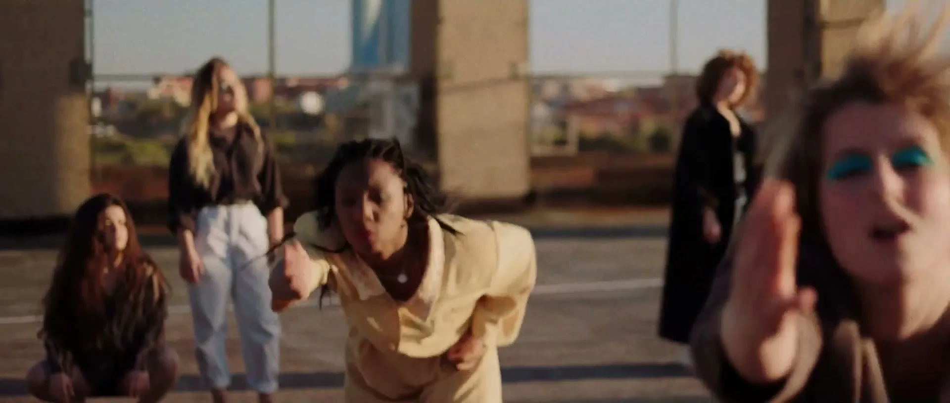 A diverse group of young women dance energetically on a concrete rooftop at golden hour, with a city skyline in the background.