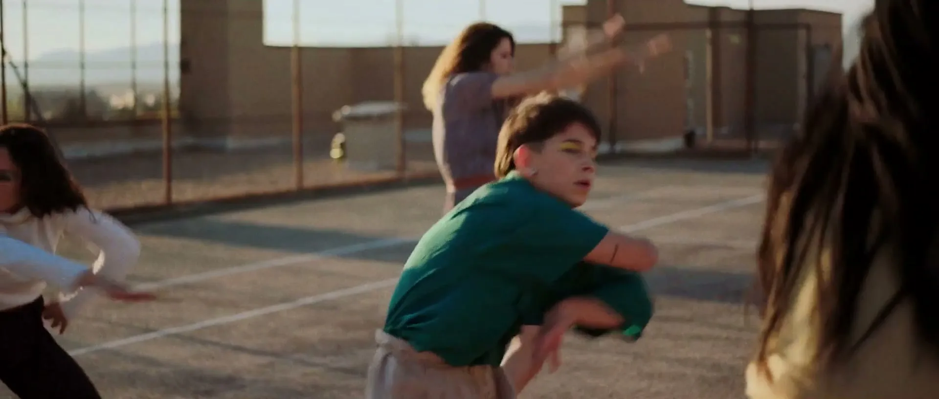 A diverse group of young women in casual attire dance with expressive movements on an outdoor rooftop court during golden hour.