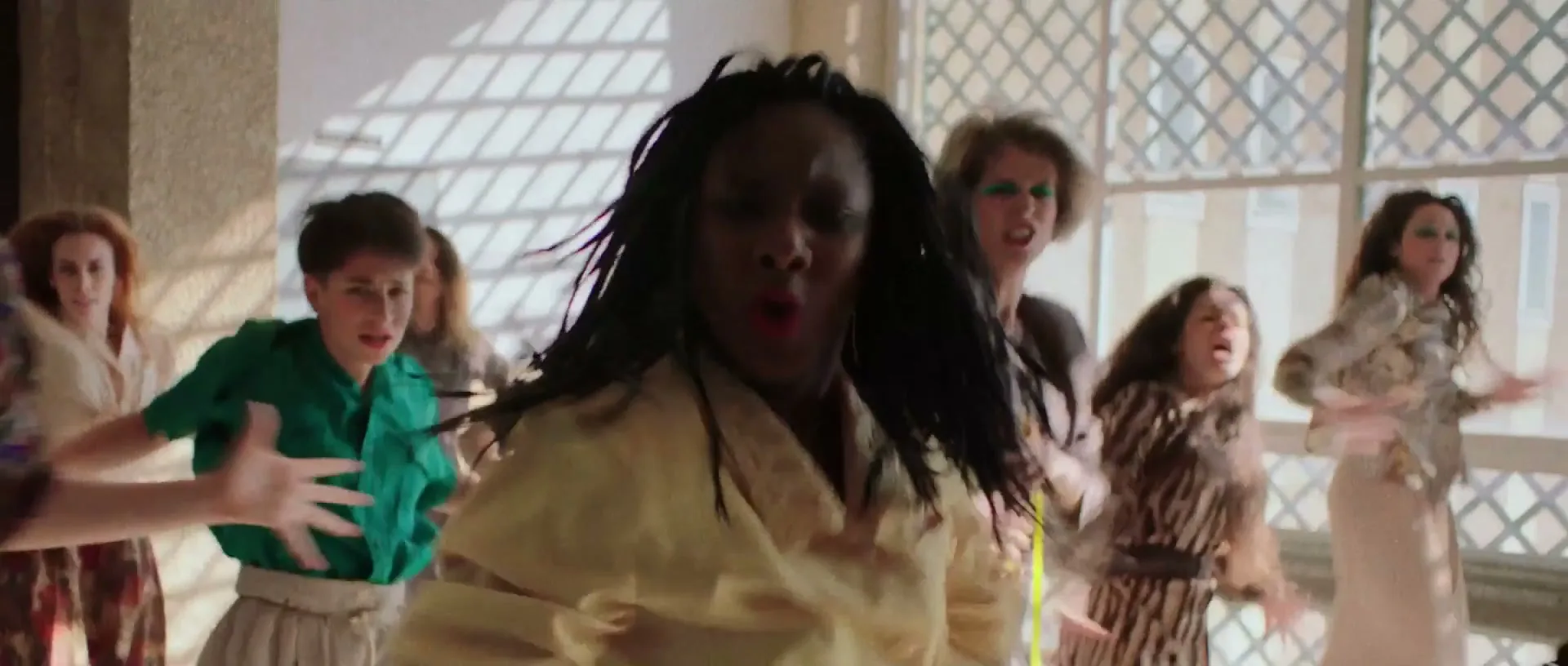 A diverse group of women in 80s-inspired outfits dance energetically in a sunlit room with large windows.
