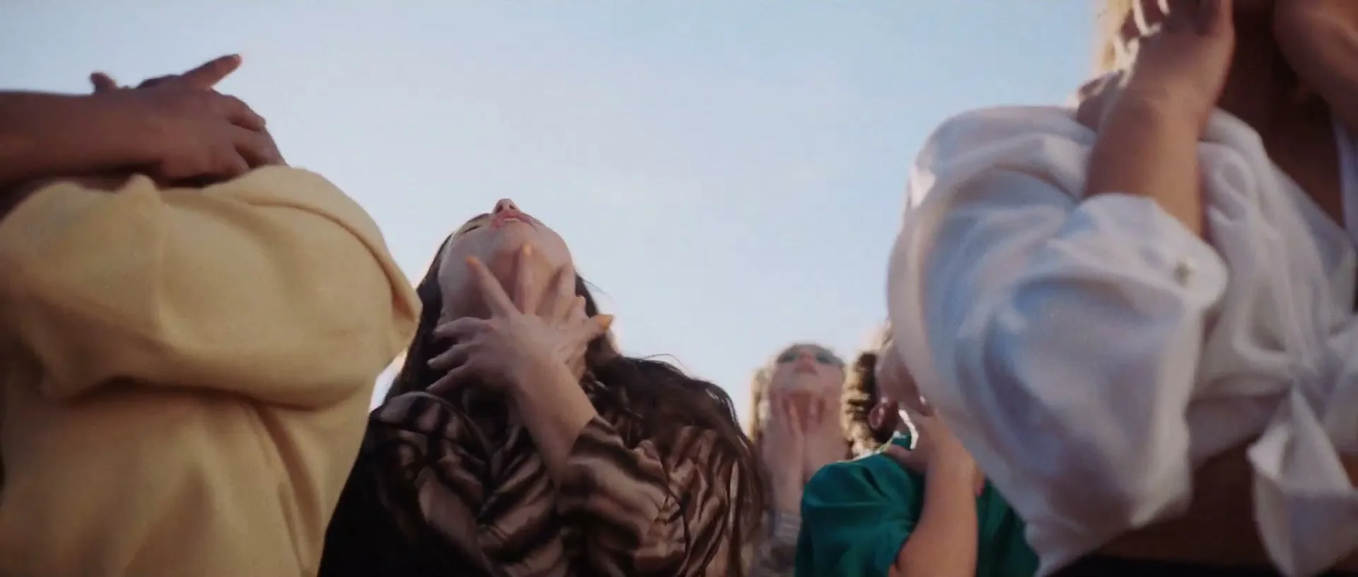 A group of young women, dressed in casual attire, dance with their heads tilted back and hands around their necks under a bright sky.