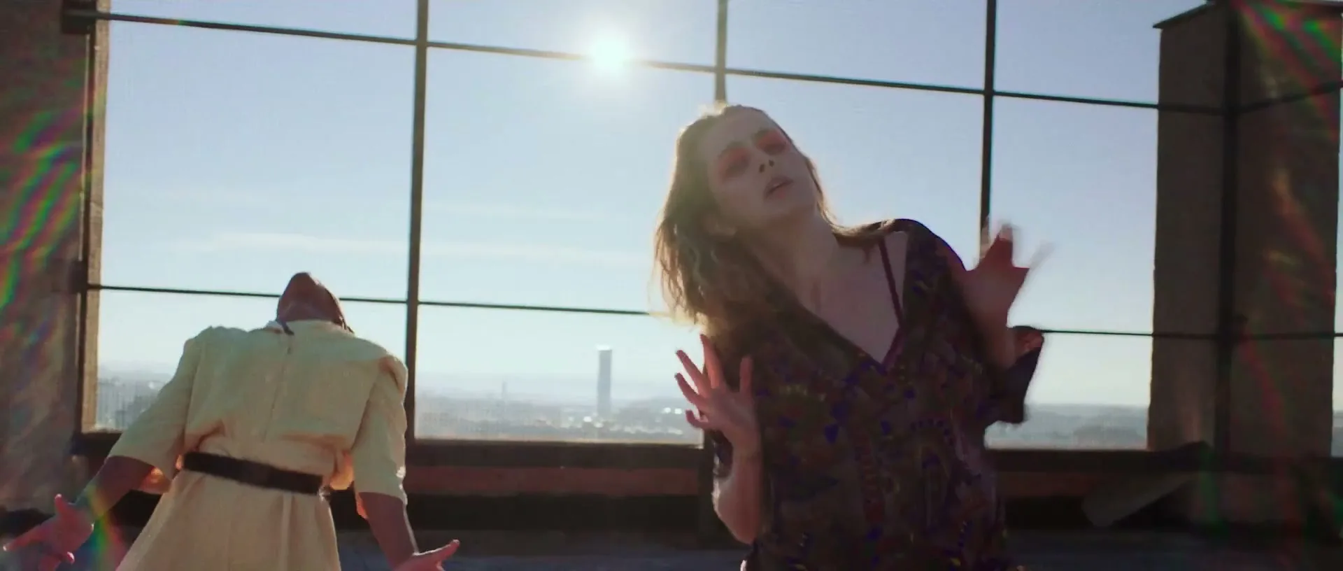 Two young women dance expressively on a sunlit rooftop with a city skyline in the background, moving their arms and bodies.