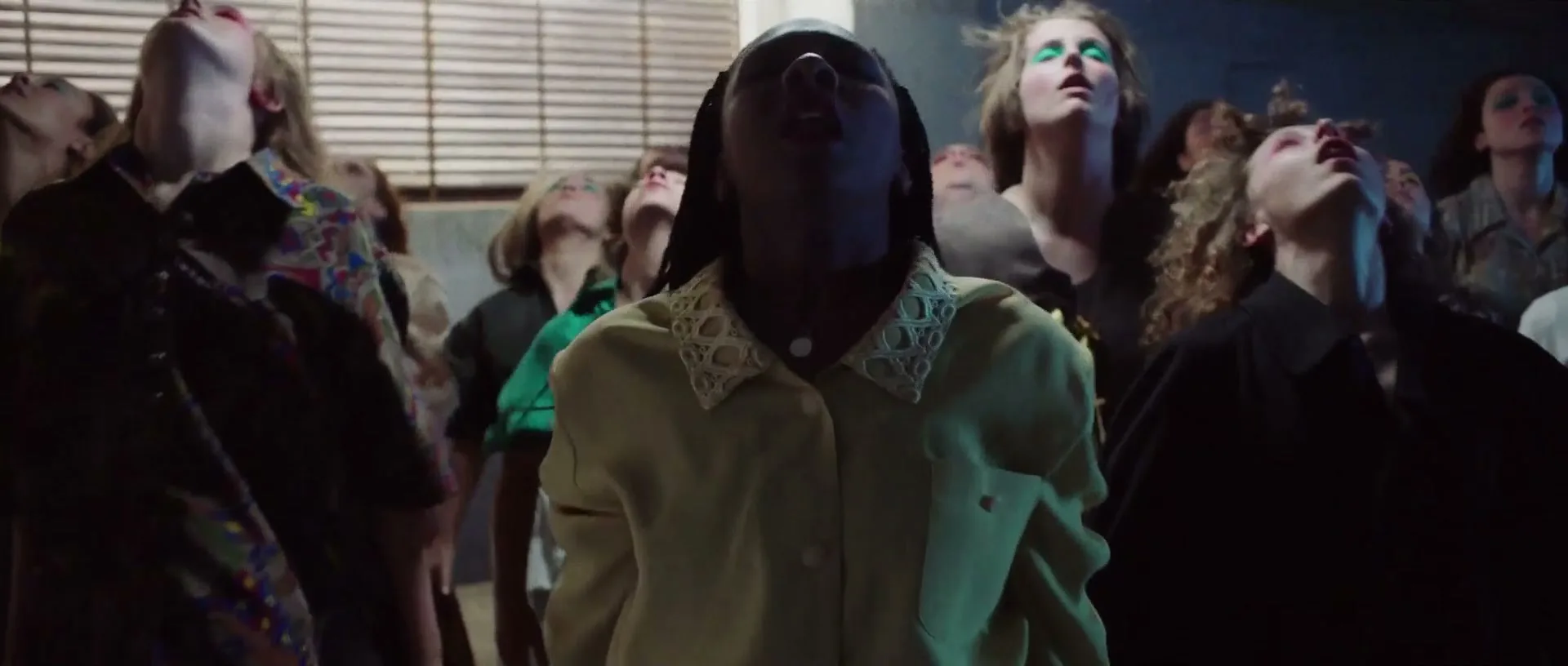 A diverse group of young people, dressed in various casual outfits, slowly rise from a bowed position, looking upwards with open mouths in a dimly lit concrete room.