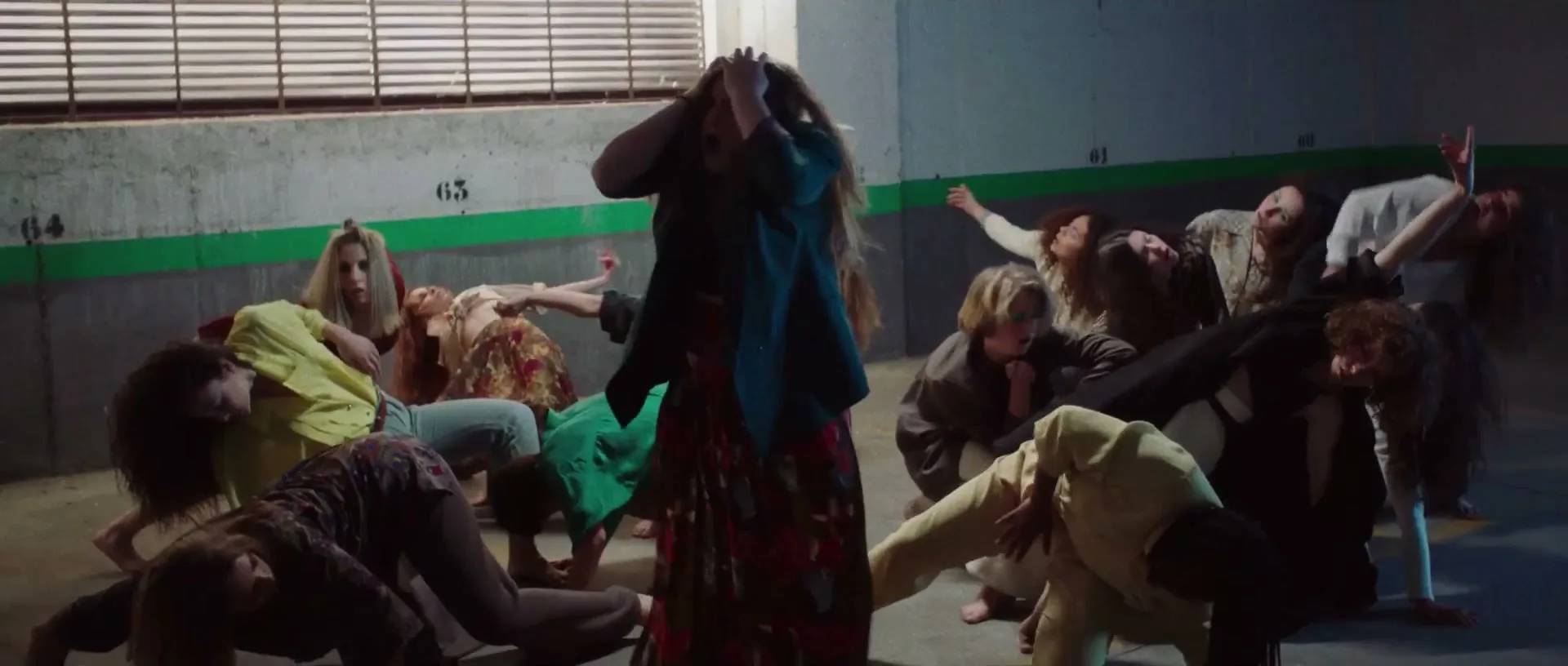 A group of women in various casual outfits are dancing in a dimly lit underground parking garage, with one woman in the center holding her head.