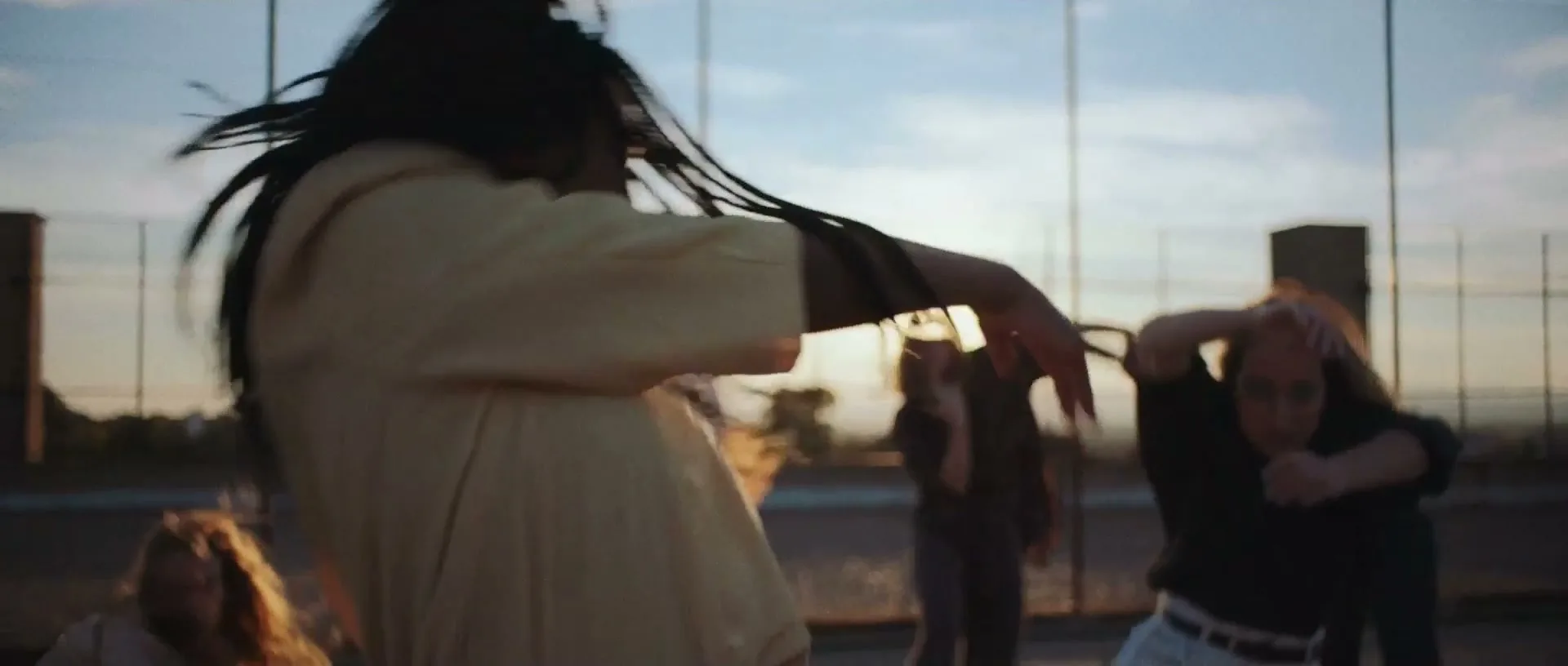 A diverse group of young people dance energetically on a rooftop at sunset, with one person in the foreground shaking their dreadlocks.